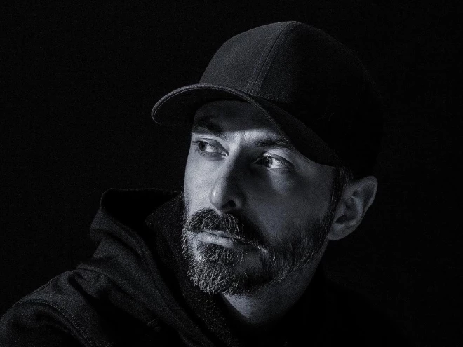 Retrato em preto e branco de um homem de barba e boné preto, vestido com moletom escuro, olhando para o lado. A iluminação destaca os traços do rosto em um fundo preto, criando uma composição de forte contraste.
