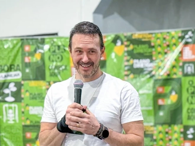 Vinicius De Tomasi Ribeiro aparece segurando um microfone durante uma apresentação. Ele veste camiseta branca, usa relógio e pulseiras, e está diante de um painel com elementos gráficos predominantemente verdes.