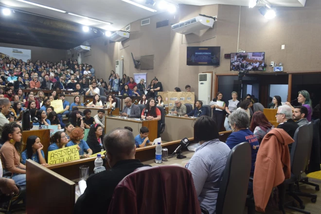 Reunião pública na Câmara de Vereadores de Caxias do Sul discute os atrasos nos repasses à assistência social. O plenário está lotado, com representantes de entidades e cartazes cobrando recursos para manter os atendimentos.