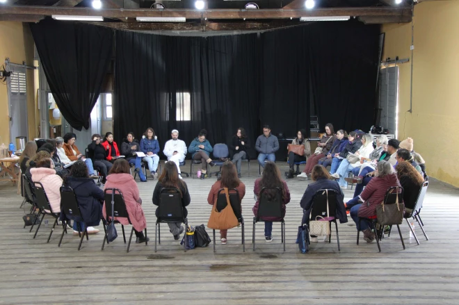 Grupo de professores participa de encontro de formação em Caxias do Sul. Os participantes estão sentados em círculo, em um espaço com piso de madeira e palco ao fundo.