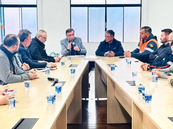 Reunião do Gabinete de Crise reúne autoridades e integrantes de equipes de emergência em torno de uma mesa, com documentos, celulares e copos de água. Um dos participantes fala ao grupo usando um microfone.