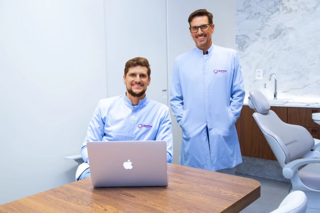 Dois profissionais de odontologia posam em uma clínica. Um deles está sentado diante de um notebook e o outro permanece em pé; ambos usam uniformes da Benne Odonto.