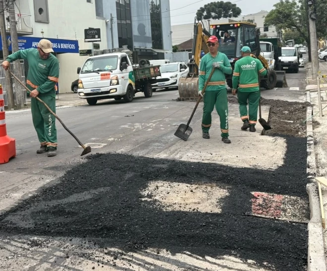 Funcionários da Codeca realizam reparos no pavimento de uma via em Caxias do Sul, com aplicação de asfalto em trecho danificado. Uma retroescavadeira e veículos de serviço acompanham a operação, que ocupa parte da pista.