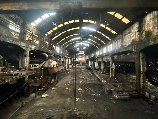 Interior do bloco 1 da Maesa, com estrutura industrial antiga, paredes de tijolos, pilares, máquinas e instalações desgastadas. A cobertura metálica apresenta aberturas que permitem a entrada de luz natural.