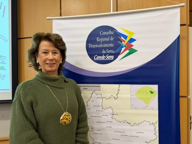 Presidente do Corede Serra, Monica Mattia, posa diante de um painel com o mapa dos municípios da região e a identificação do Conselho Regional de Desenvolvimento da Serra. Ela veste blusa verde e colar com pingente decorativo.