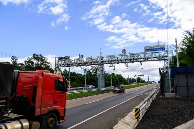 Foto mostra uma rodovia com pórtico de pedágio eletrônico da CSG. Um caminhão vermelho e um carro trafegam pela pista sob o equipamento, com sinalização de limite de 80 km/h e indicação de pedágio eletrônico.