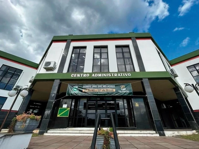 Fachada do Centro Administrativo da Prefeitura de São Marcos, com entrada principal envidraçada, paredes brancas e detalhes em verde e preto. Na frente do prédio, há um banner institucional, vasos com plantas e luminárias externas.