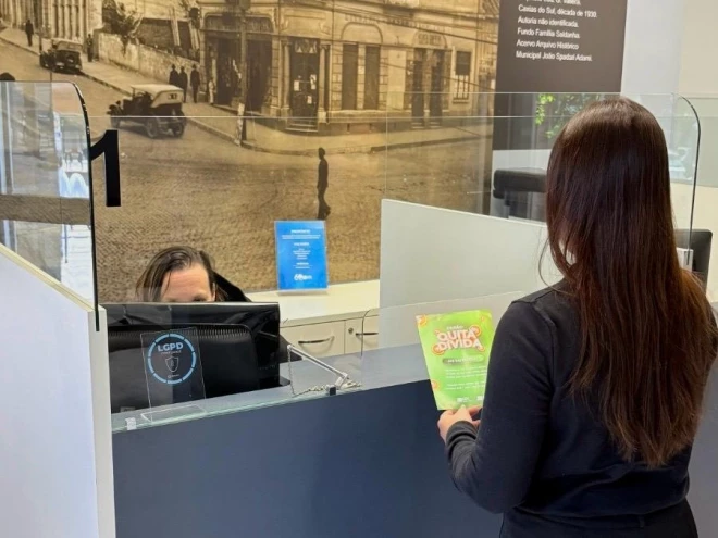Mulher apresenta material informativo do Feirão Quita Dívida no balcão de atendimento da CDL Caxias. Ao fundo, uma atendente está diante de um computador, em ambiente com painéis históricos de Caxias do Sul.