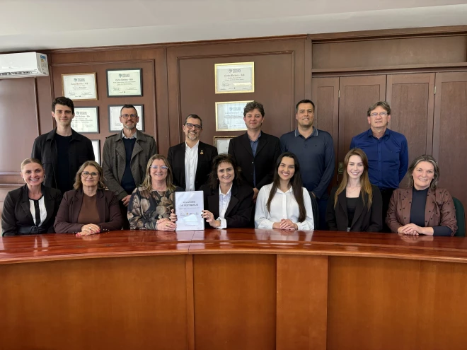 Grupo de pessoas posa para foto em uma sala, atrás de uma mesa de madeira. Uma mulher ao centro segura um relatório do 34º FestiQueijo.