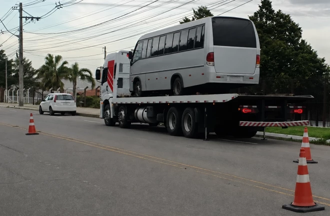Ônibus de fretamento é transportado por um caminhão-guincho durante operação de fiscalização em uma via urbana de Caxias do Sul. Cones sinalizam o local, enquanto veículos circulam pela rua.