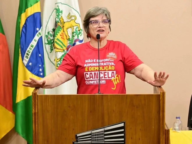 A presidente do Sindiserv Silvana Piroli discursa na tribuna da Câmara de Vereadores de Caxias do Sul. Ela veste camiseta vermelha com mensagem de mobilização sindical. Ao fundo, aparecem as bandeiras do Brasil, do Rio Grande do Sul e de Caxias do Sul.