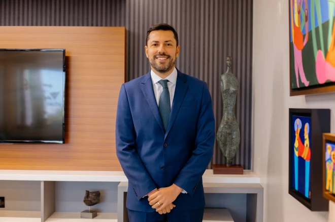 em Pauta em até 250 Gustavo Neves Rocha posa para foto em ambiente interno, usando terno azul, camisa branca e gravata estampada. Ao fundo, há uma televisão, esculturas decorativas e quadros coloridos em uma parede com detalhes em madeira.