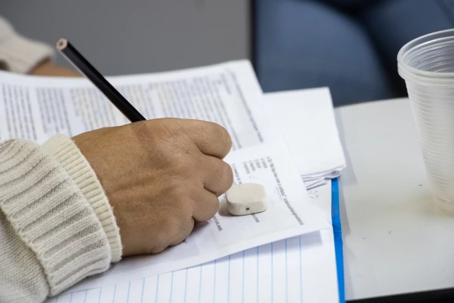 Mão de estudante escreve com lápis sobre uma folha de atividade escolar, apoiada em um caderno. Sobre a mesa, há uma borracha, um lápis e um copo plástico.