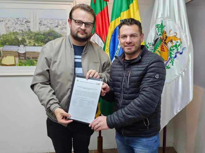 Vereadores Wagner Petrini e João Uez seguram documento durante a transmissão temporária da Presidência da Câmara de Caxias do Sul, com bandeiras oficiais ao fundo.