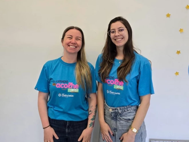 Duas mulheres sorriem para a foto usando camisetas azuis do projeto “Um legado que acolhe e ensina”, da instituição Madre Imilda. Ao fundo, há uma parede clara decorada com pequenas estrelas amarelas.