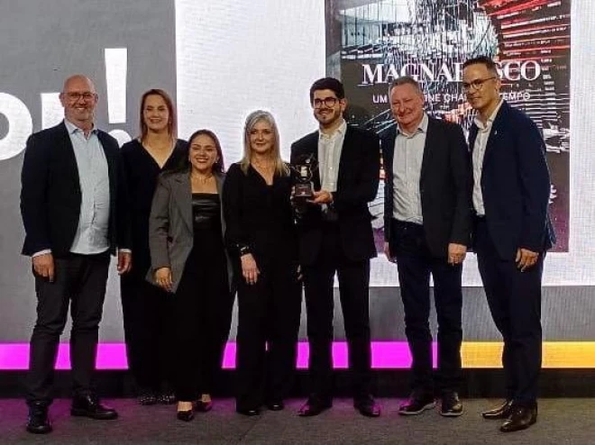 Foto mostra sete pessoas em um palco durante uma premiação. O grupo aparece lado a lado, sorrindo, com um troféu ao centro. Ao fundo, um telão exibe a capa do livro “Magnabosco: Um Magazine Chamado Tempo”.