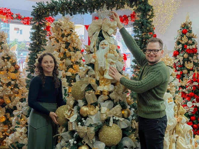 Sócios da Sunflower Home & Decor posam ao lado de árvores de Natal decoradas com laços dourados, bolas e enfeites temáticos no showroom inaugurado em Caxias do Sul.
