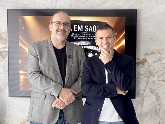 Os médicos André Leite e Vinícius Lain posam diante de uma tela que exibe a capa de um livro sobre inteligência artificial e saúde.