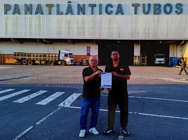 Dois homens seguram um documento em frente à unidade industrial da Panatlântica Tubos, em Caxias do Sul. Ambos vestem camisetas pretas, enquanto caminhões estão estacionados na área de acesso à fábrica.