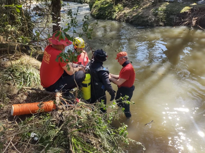 Equipe de resgate realiza buscas em um curso d’água, com um mergulhador equipado para entrar na água. Outros profissionais auxiliam a operação às margens, cercadas por vegetação e galhos.