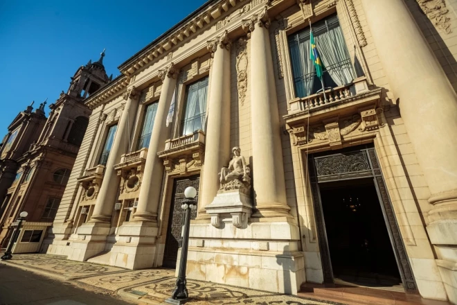 Fachada do Palácio Piratini, sede do governo do Rio Grande do Sul, em Porto Alegre. O prédio histórico apresenta colunas ornamentadas, esculturas, grandes janelas e uma bandeira do Brasil na fachada, sob céu azul.