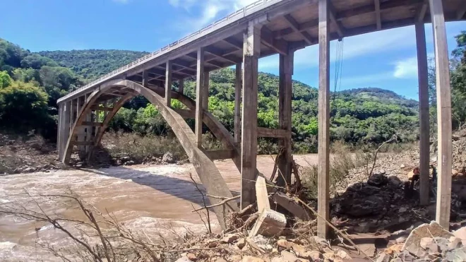 Ponte sobre o Rio das Antas apresenta pilares e arcos de concreto, com trechos da estrutura cercados por galhos e pedras. Abaixo, o rio corre entre áreas de vegetação e encostas cobertas por mata.