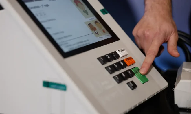 Urna eletrônica em close, com a tela de votação e o teclado numérico. Uma mão pressiona a tecla verde “CONFIRMA”, enquanto os botões “CORRIGE” e “BRANCO” aparecem no equipamento.
