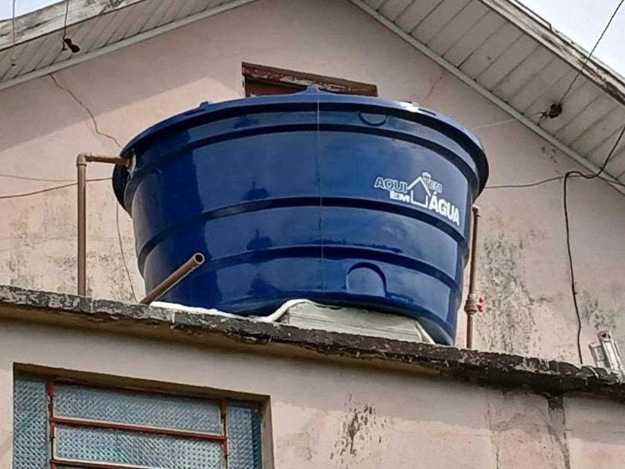 em Pauta em até 250 Caixa-d’água azul do programa Aqui em Casa Tem Água instalada sobre a laje de uma residência, junto ao telhado e às tubulações do imóvel.