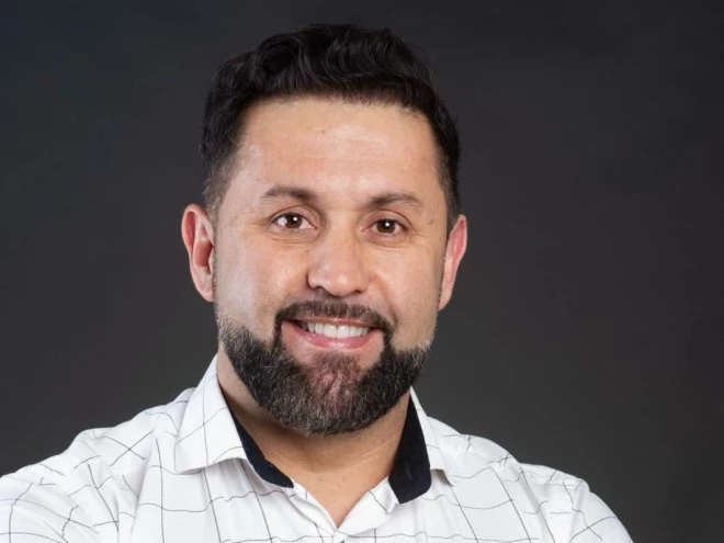Leno Almeida, diretor de Negócios e Gestão da Saúde do Círculo Saúde de cabelos escuros e barba aparece sorrindo em retrato formal, usando camisa branca com detalhes em preto, diante de um fundo escuro.