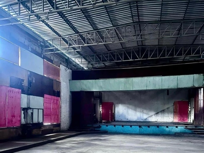 Interior do prédio histórico de Galópolis, em Caxias do Sul, com estrutura metálica aparente no telhado, paredes desgastadas e palco ao fundo. O espaço integra o projeto de restauro do antigo imóvel do Círculo Operário Ismael Chaves Barcelos.