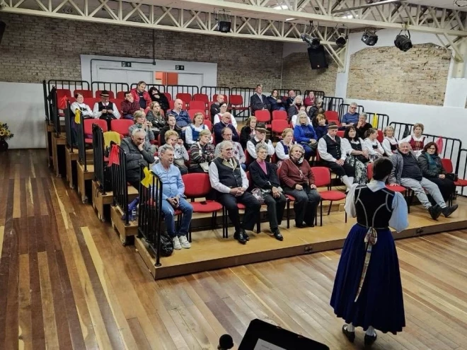 Público acompanha apresentação cultural durante a Semana das Etnias, em Caxias do Sul. Mulher vestida com traje típico italiano se apresenta diante de espectadores acomodados em arquibancadas no espaço cultural.