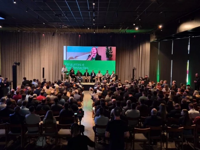 Público acompanha evento em auditório, com participantes sentados diante de um palco. Um painel de projeção exibe a imagem de uma mulher e informações sobre o encontro, enquanto convidados permanecem sentados no palco.