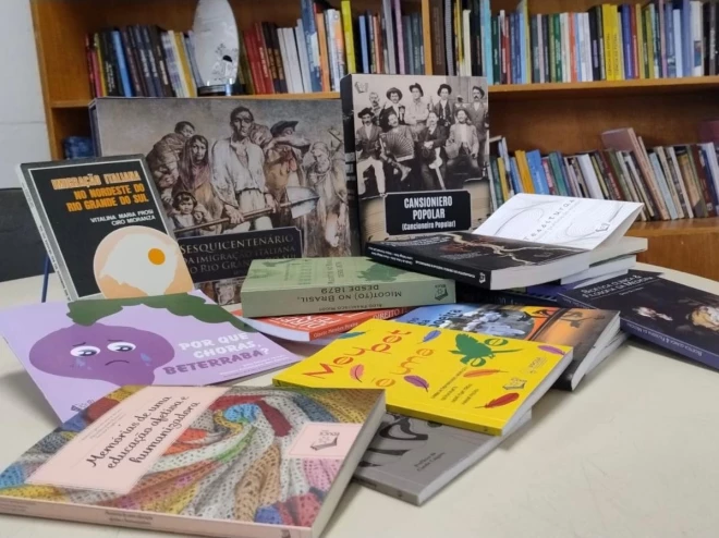 Fotografia mostra livros da EDUCS dispostos sobre uma mesa, com uma estante repleta de publicações ao fundo, destacando a diversidade do acervo editorial da Universidade de Caxias do Sul.