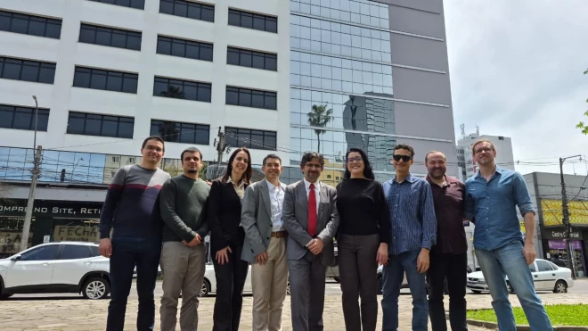 Grupo de nove pessoas posa em frente a um edifício comercial envidraçado, em Caxias do Sul. Os integrantes estão alinhados na calçada, vestindo roupas sociais e casuais, diante de uma avenida com veículos estacionados.