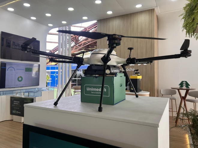 Drone da Unimed Serra Gaúcha exposto sobre uma bancada durante a Mercopar, em Caxias do Sul. O equipamento possui hélices e compartimento verde para transporte de amostras laboratoriais.